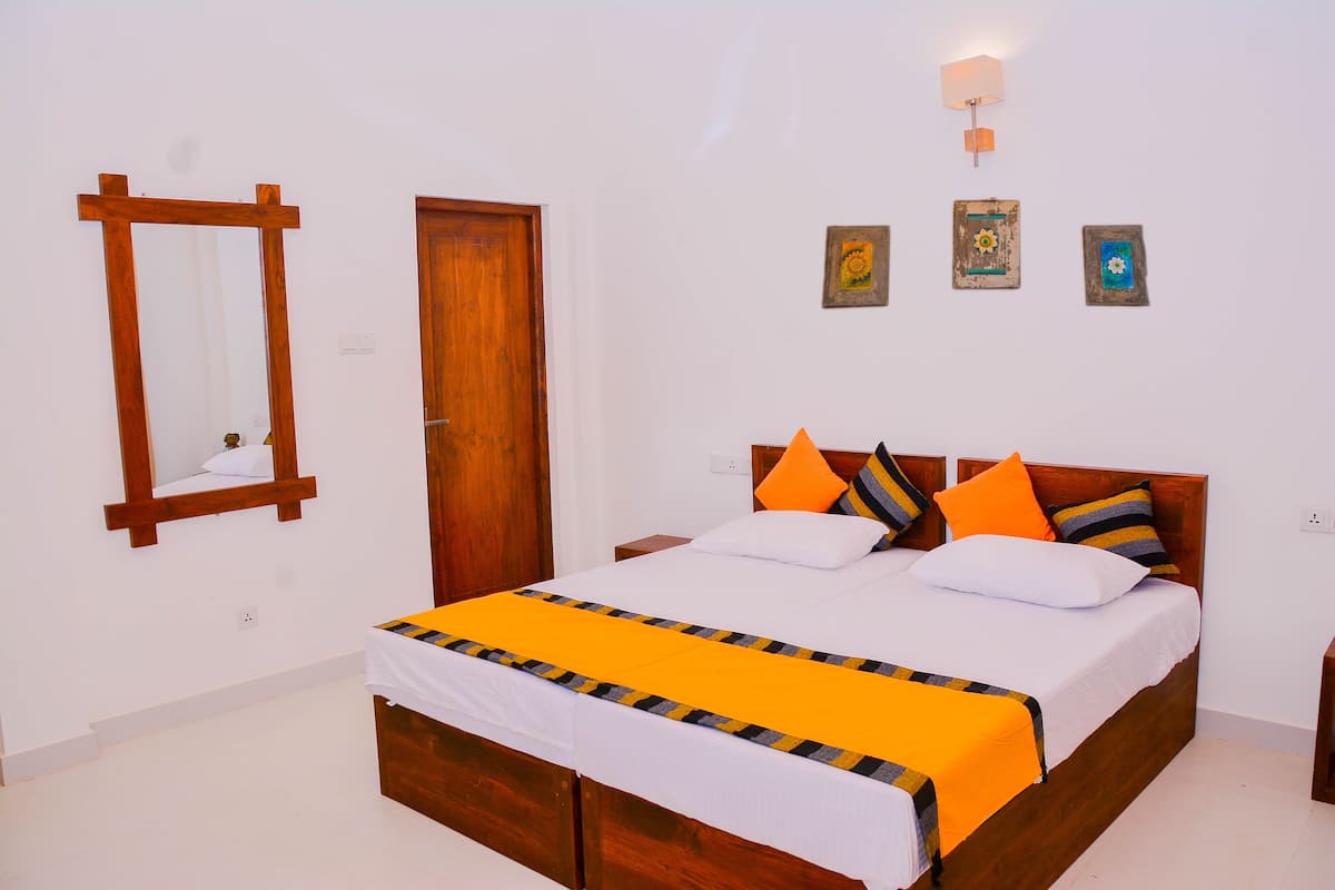 Quarto com cama de madeira, roupa de cama branca e manta listrada em amarelo e preto. Há uma porta de madeira e um espelho fixado na parede.