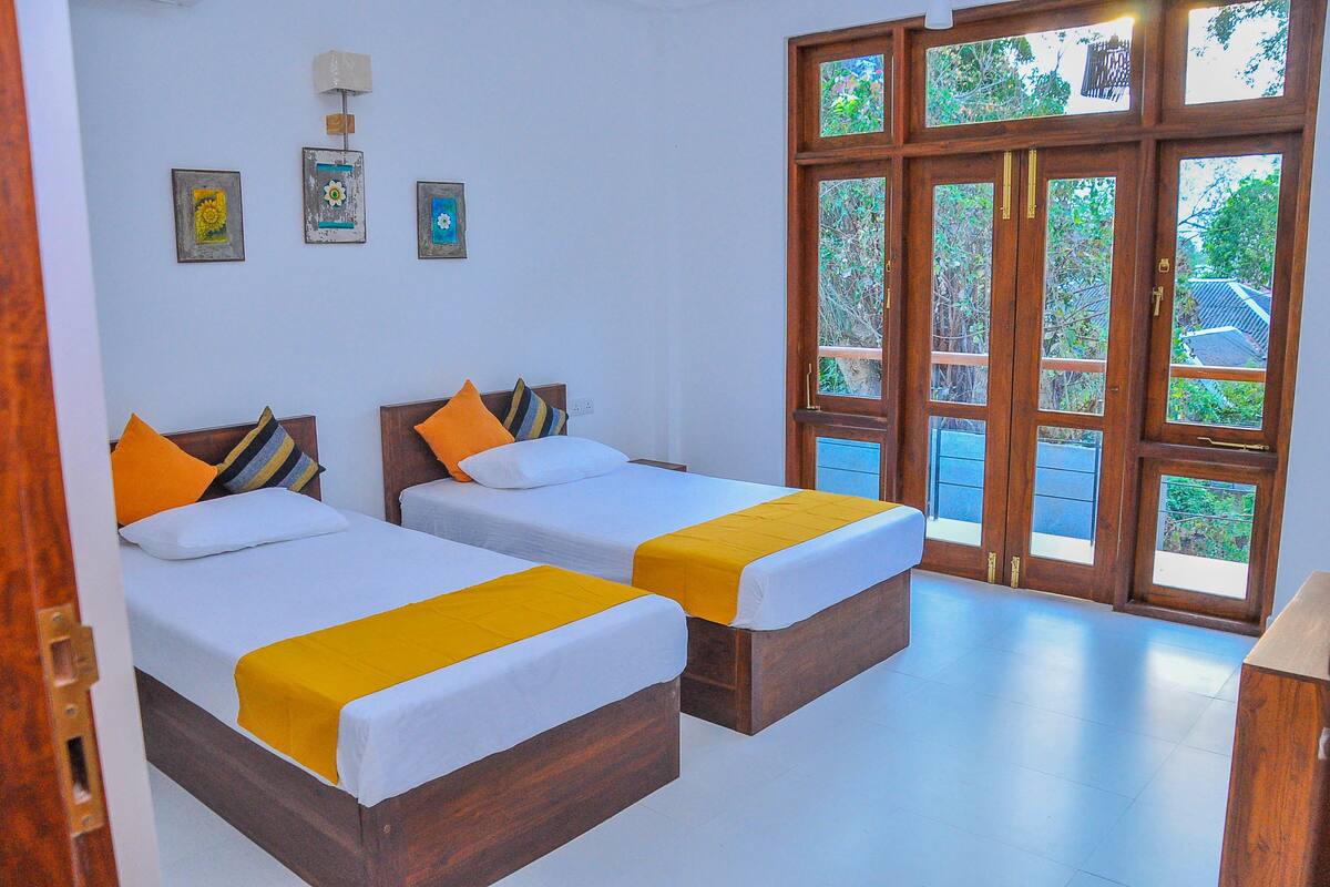 Quarto com duas camas, cada uma com colcha amarela, roupa de cama branca e almofadas laranja. Há quadros nas paredes e uma janela grande com vista para o verde.