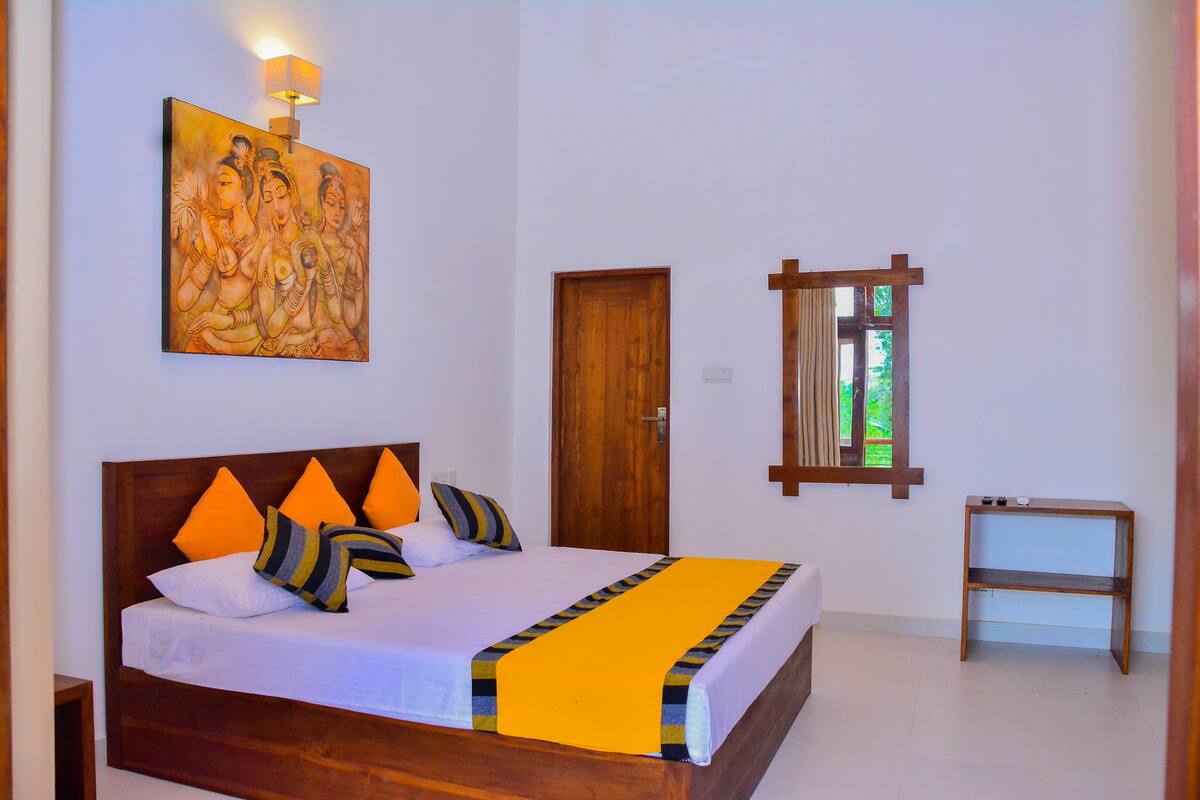 Quarto com cama de madeira, almofadas laranja, colcha branca, manta amarela, porta de madeira, janela com vista e pintura pendurada na parede.