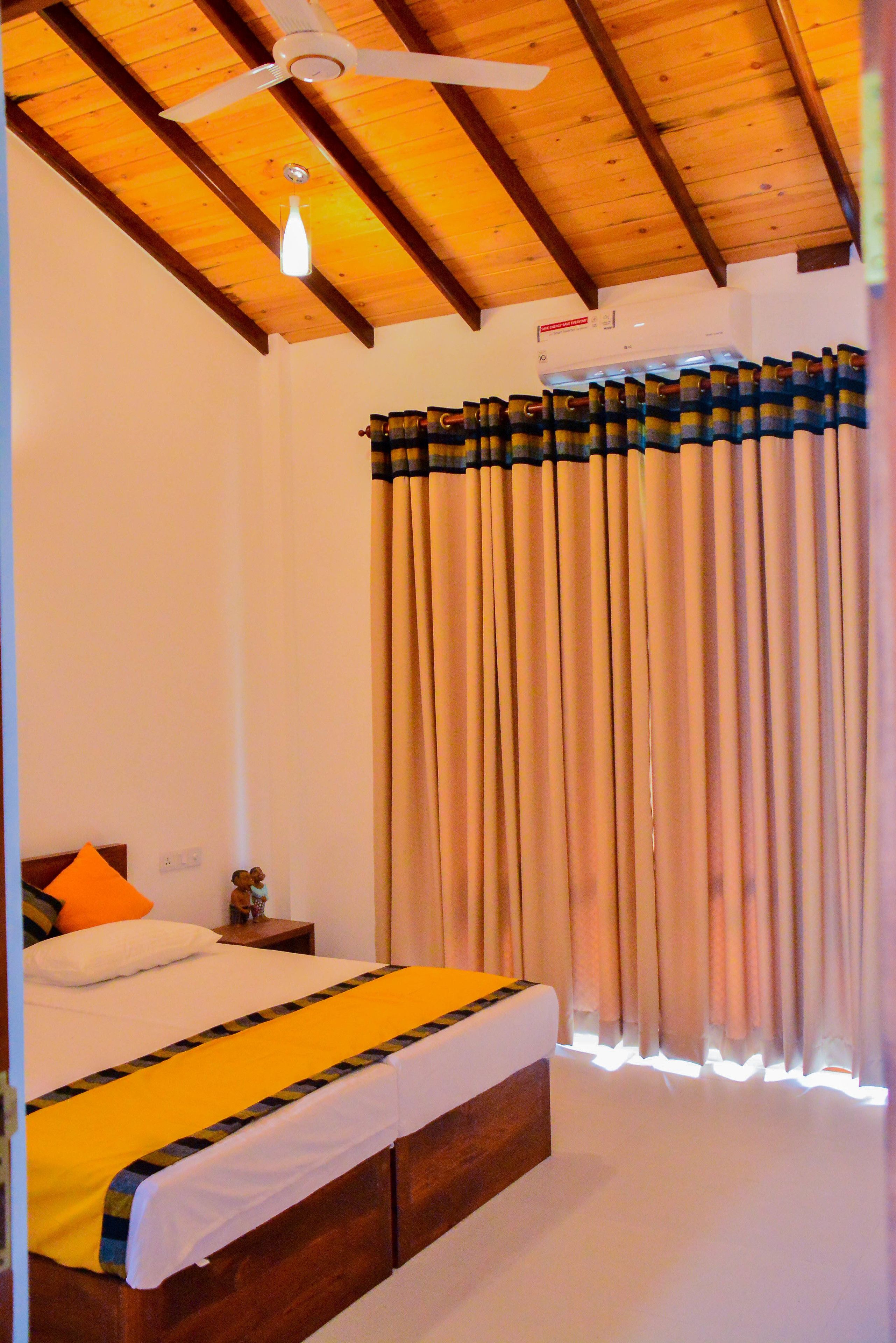 A bedroom with a wooden ceiling, a bed with a yellow and black bedspread, a nightstand with a lamp, and beige curtains.
