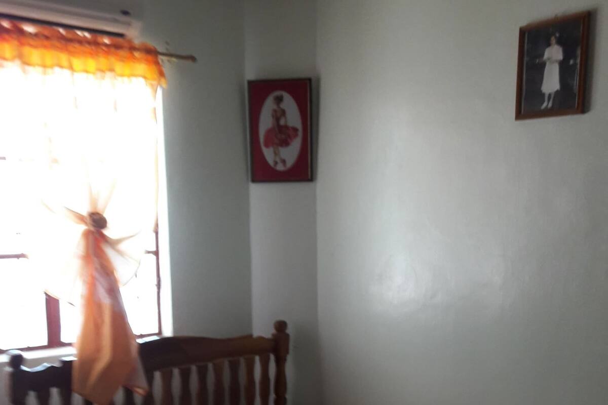 Une chambre avec un lit simple, une tête de lit en bois, une fenêtre avec des rideaux et un mur orné d’un tableau encadré.