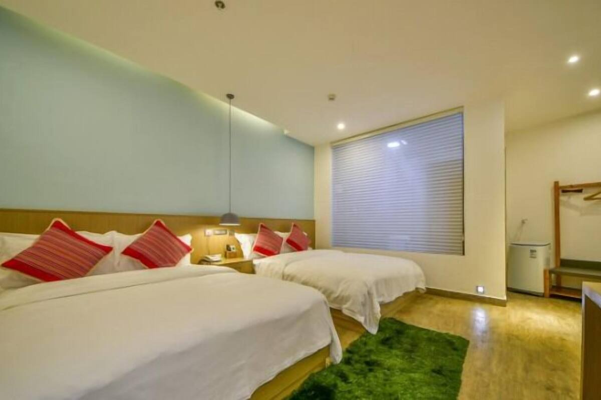 A hotel room with two beds, a green rug, a bedside table with a lamp, and a window with blinds.