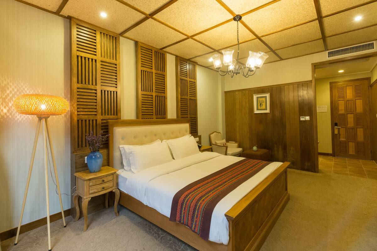 A hotel room with a large bed, wooden paneling on the walls, a chandelier, and a bedside table with a vase.