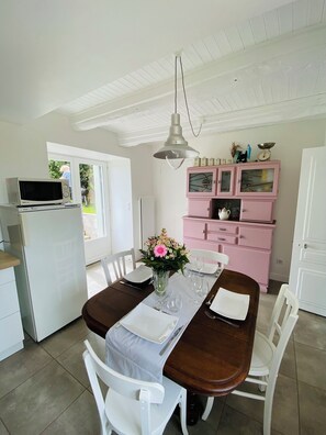 Un coin repas avec une table en bois dressée pour quatre personnes, des chaises blanches, un vase avec des fleurs et un buffet rose contenant des ustensiles de cuisine.