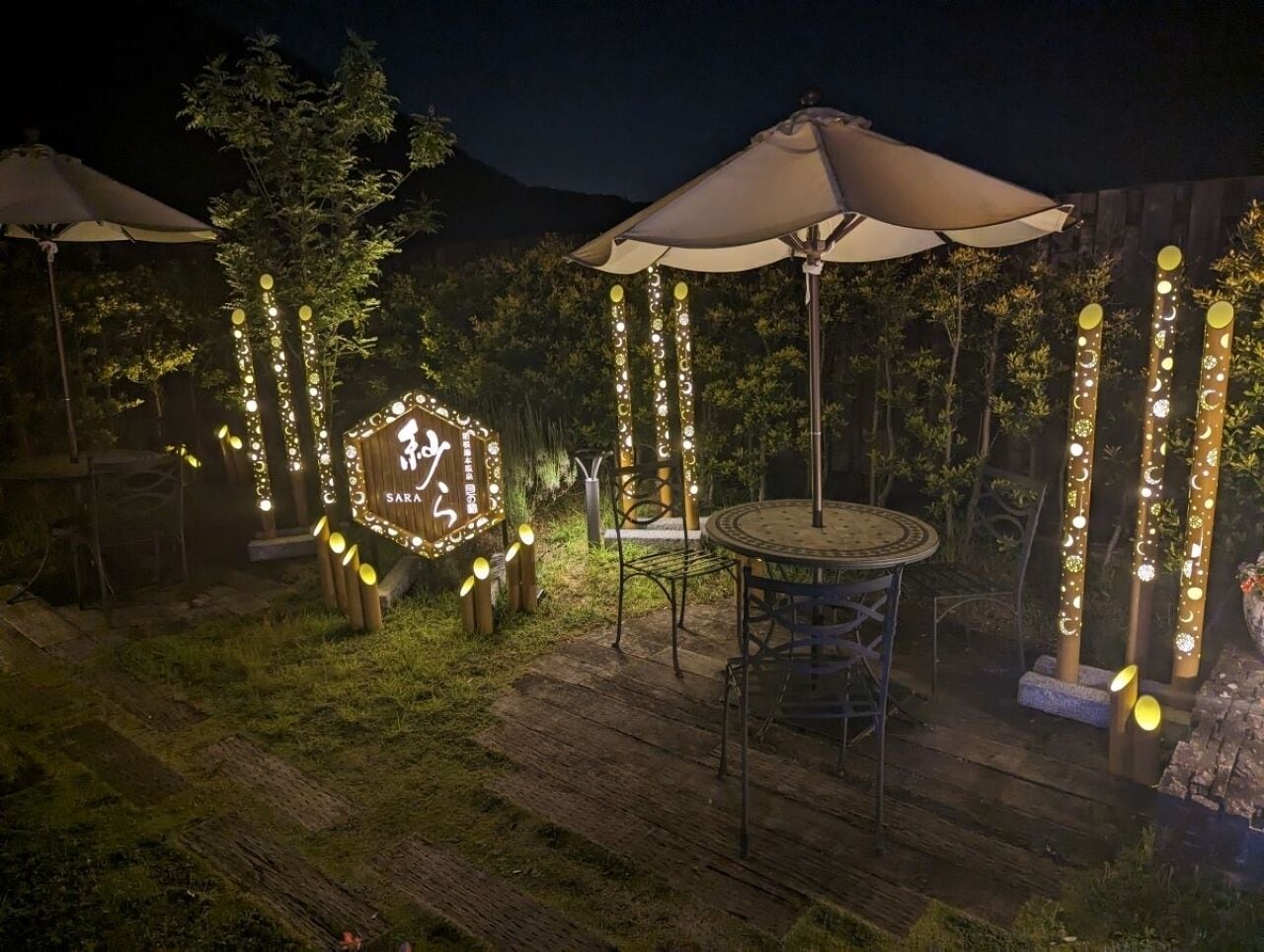 Een buitenruimte met ronde tafels en stoelen, omringd door lichtslingers en houten palen.