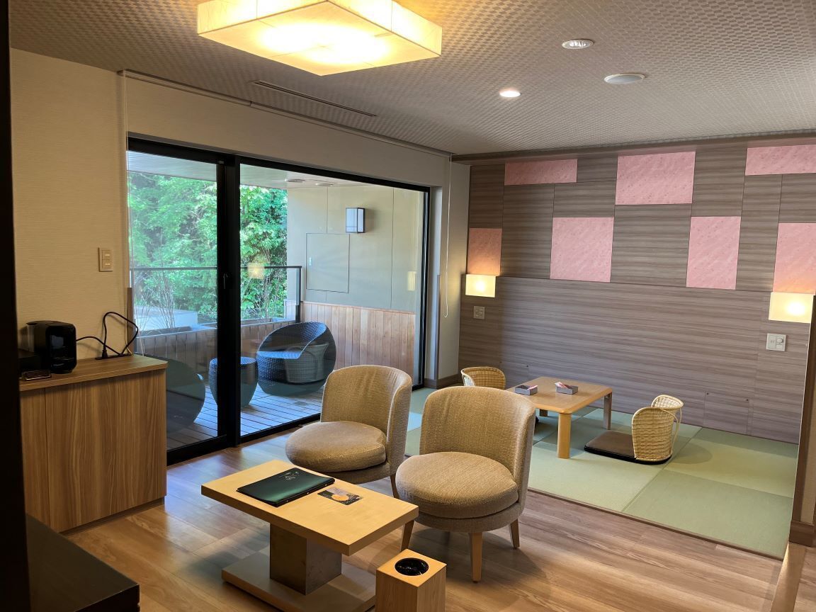 A modern hotel room with a wooden floor, a desk with a laptop, two armchairs, a small table, and a sliding glass door leading to an outdoor seating area.