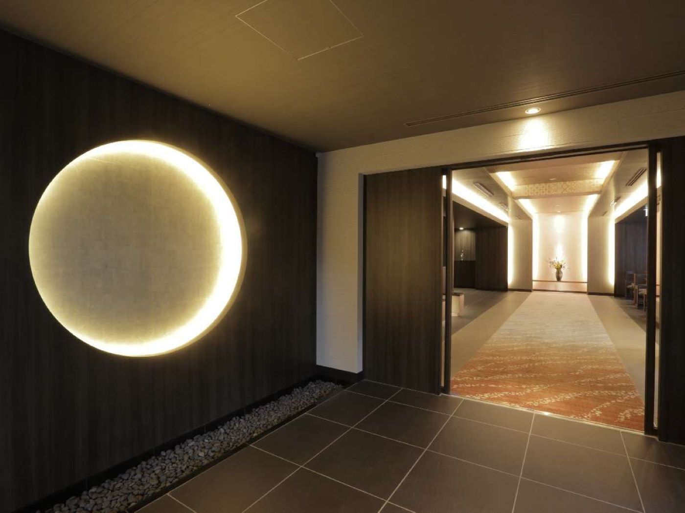 Corredor moderno de hotel com uma luminária circular na parede, piso de azulejo e área com carpete que leva a um quarto.