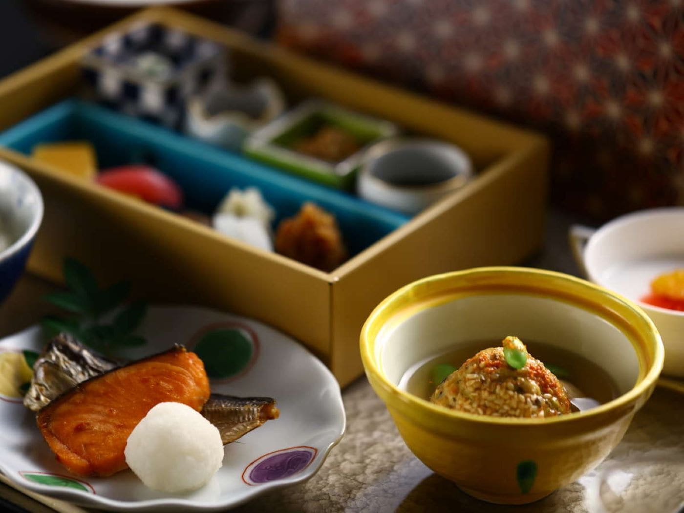 A traditional Japanese meal with a variety of dishes including salmon, rice, and pickled vegetables, served on a table with a patterned tablecloth.