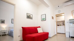 A modern living room with a red sofa, a small white table with a lamp, and a kitchen area with white cabinets and a clock on the wall.