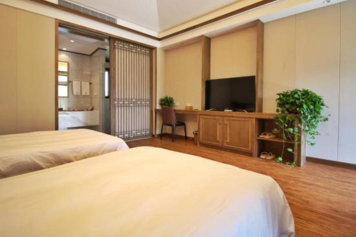 A hotel room with two beds, a desk with a TV, a plant, and a sliding door leading to another room.