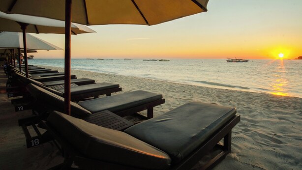 Et loungeområde ved stranden med liggestole og parasoller, med udsigt over havet i solnedgang.