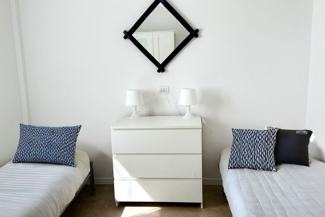 A bedroom with a white dresser, two bedside tables with lamps, a mirror, and two beds with patterned pillows.