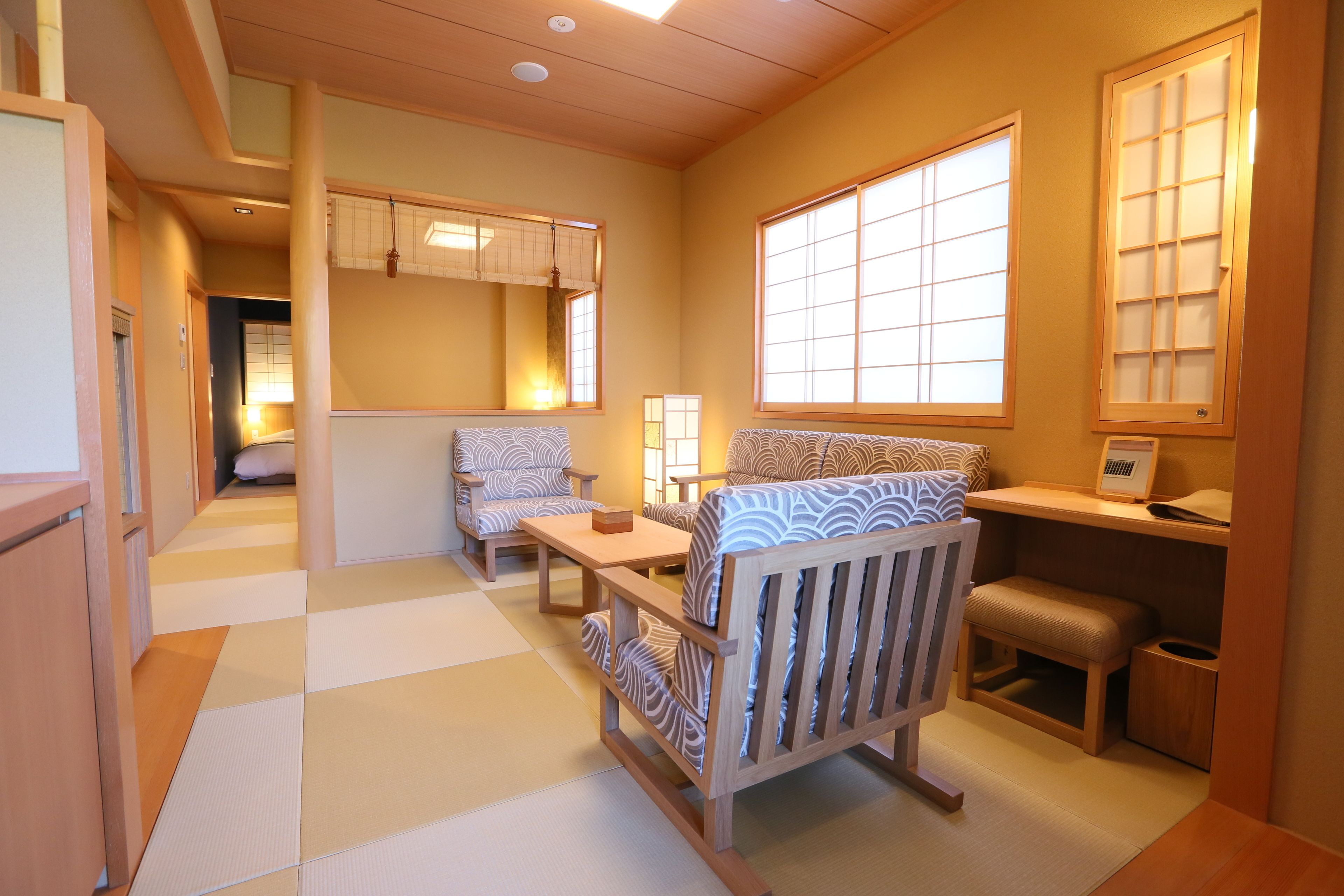 A traditional Japanese-style room with tatami flooring, a low wooden table, two patterned chairs, a small bench, and a window with a view of the outside.