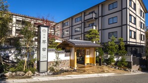 Un edificio de varios pisos con una entrada tradicional japonesa, un camino de piedra y vegetación circundante.