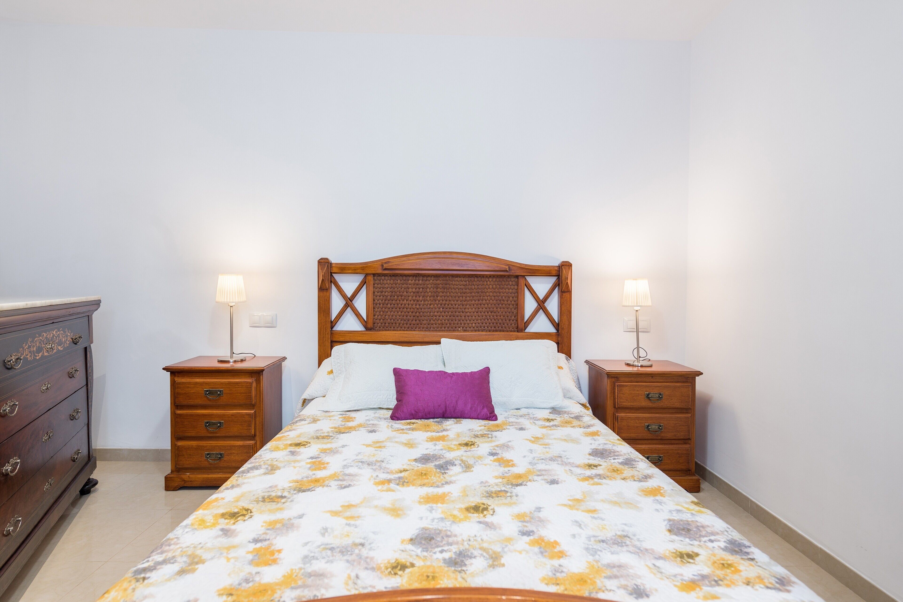 Un lit bien fait, recouvert d’une couvre-lit à motifs floraux, deux tables de chevet avec des lampes et une commode en bois.