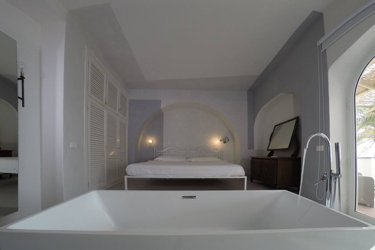 A modern bedroom with a large bathtub, a bed with a white headboard, and a view of the outdoors through a large window.