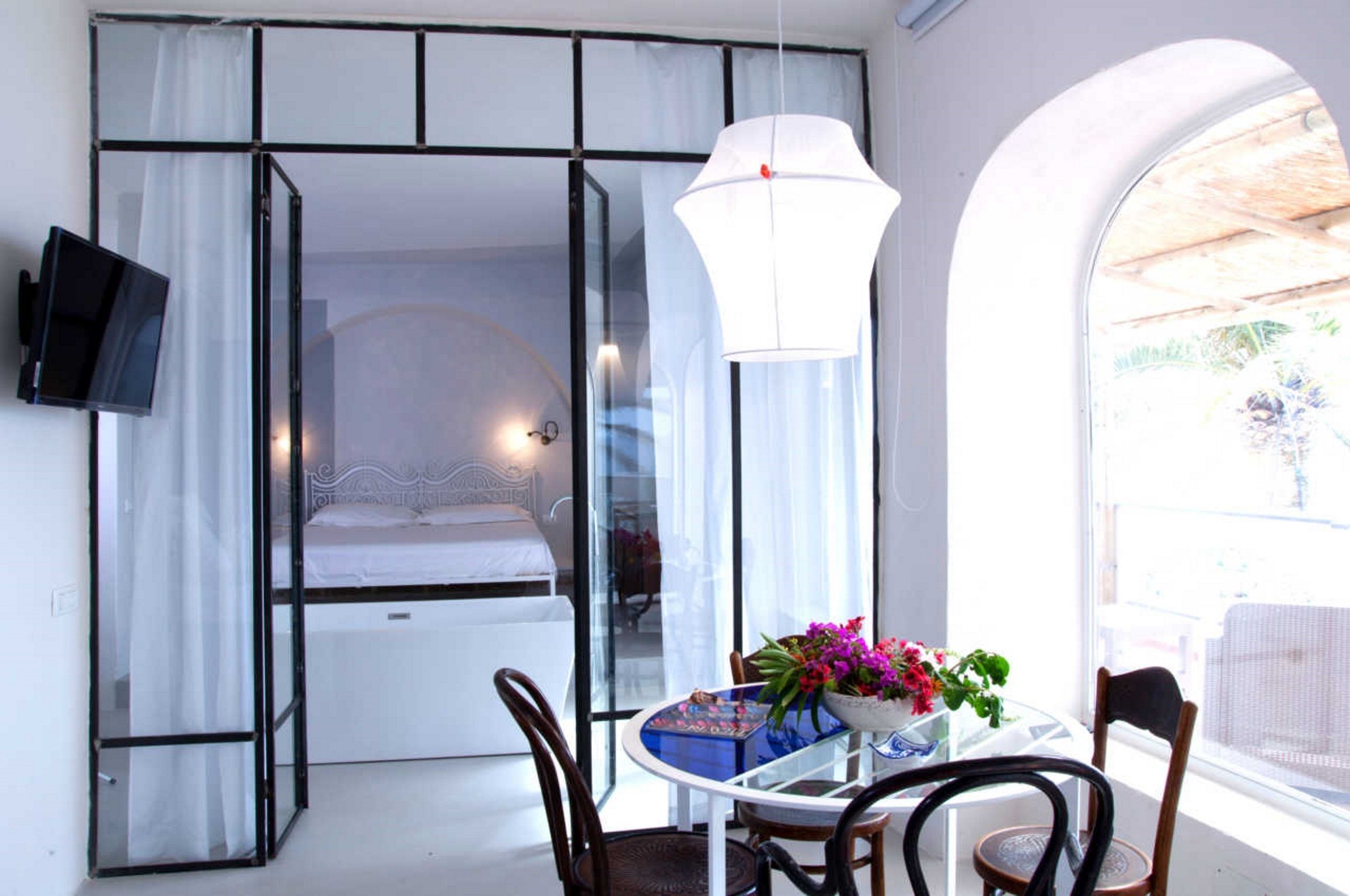 Camera d'albergo moderna con una parete divisoria in vetro che separa la zona notte dalla zona salotto. C'è una televisione montata a parete, un tavolo rotondo con sedie e un vaso con fiori.