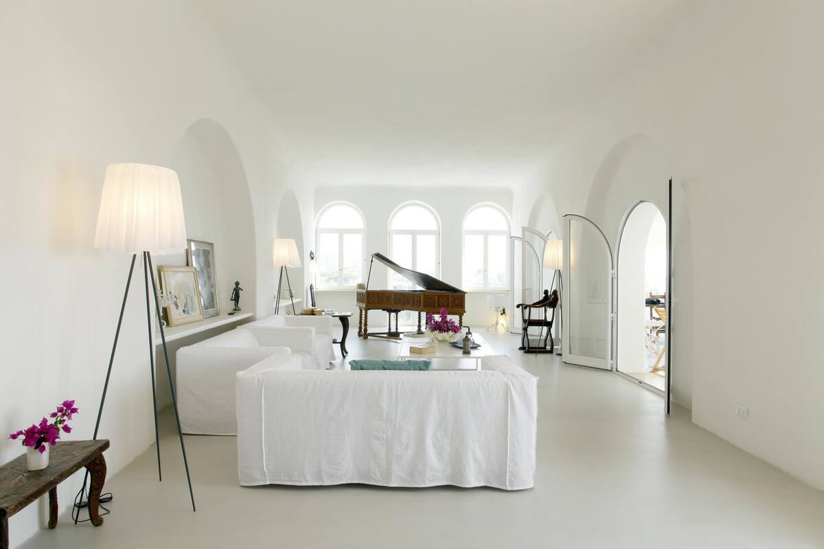 A spacious room with a grand piano, white furniture, and large arched windows.