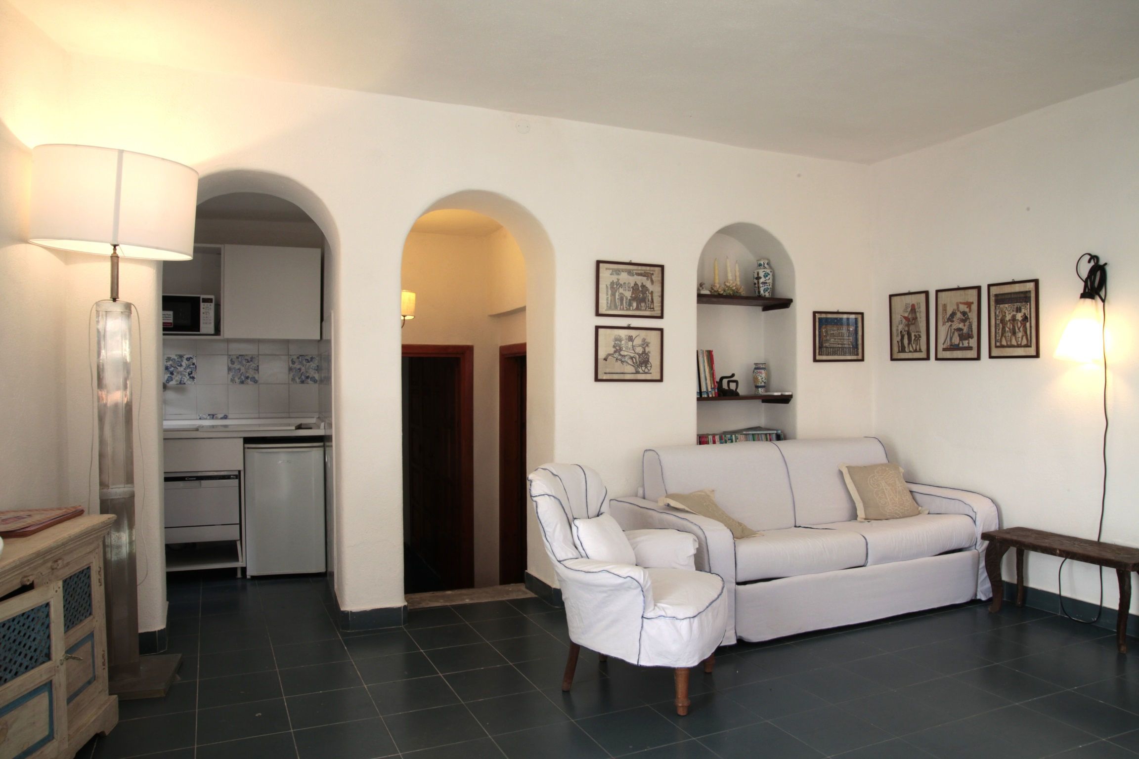 A living room with a white sofa, a white armchair, a wooden bench, and a lamp. There is a kitchen in the background with a microwave and a tiled backsplash.
