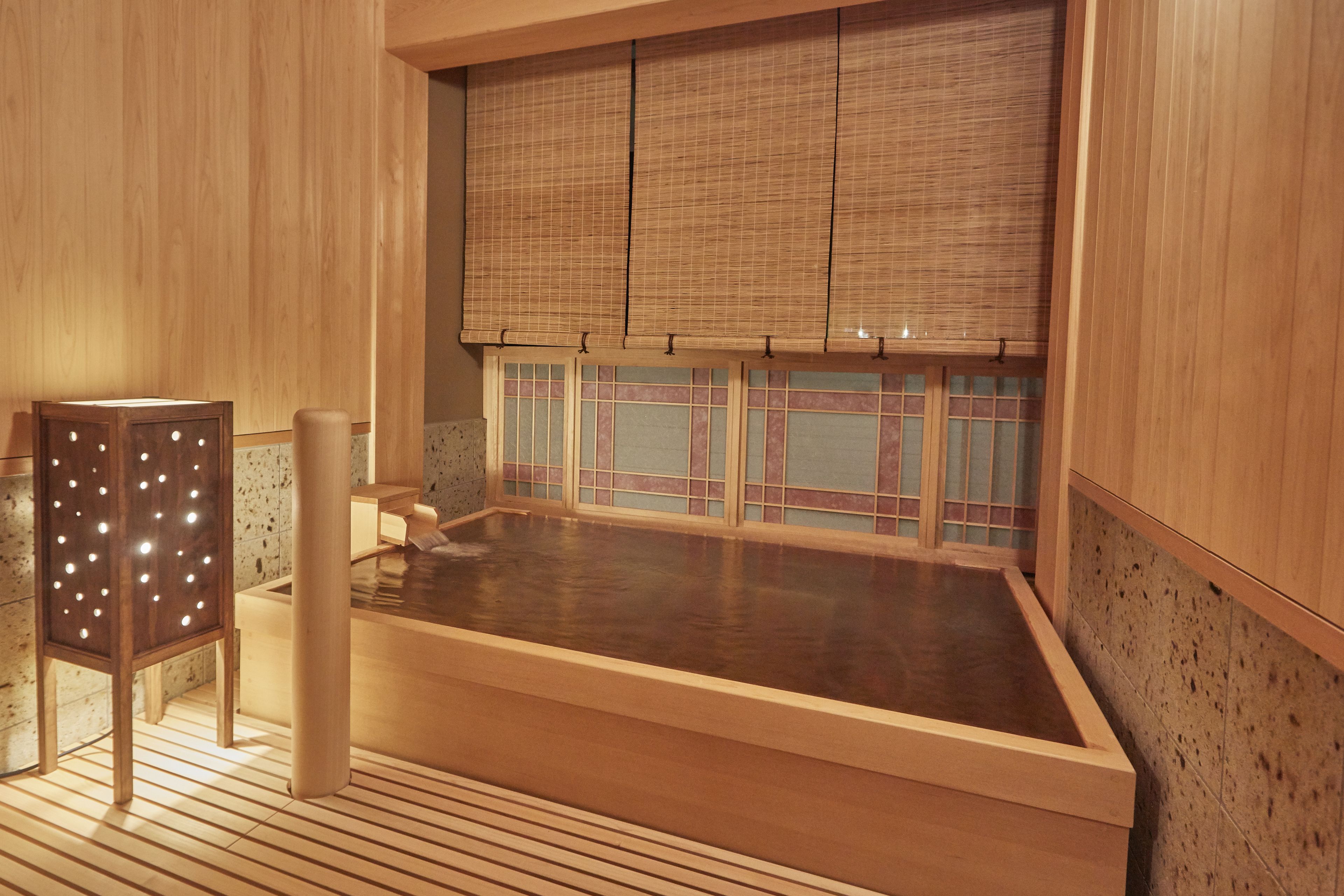 Une pièce en bois avec une grande baignoire en bois, une chaise en bois et une fenêtre avec des stores en bambou.
