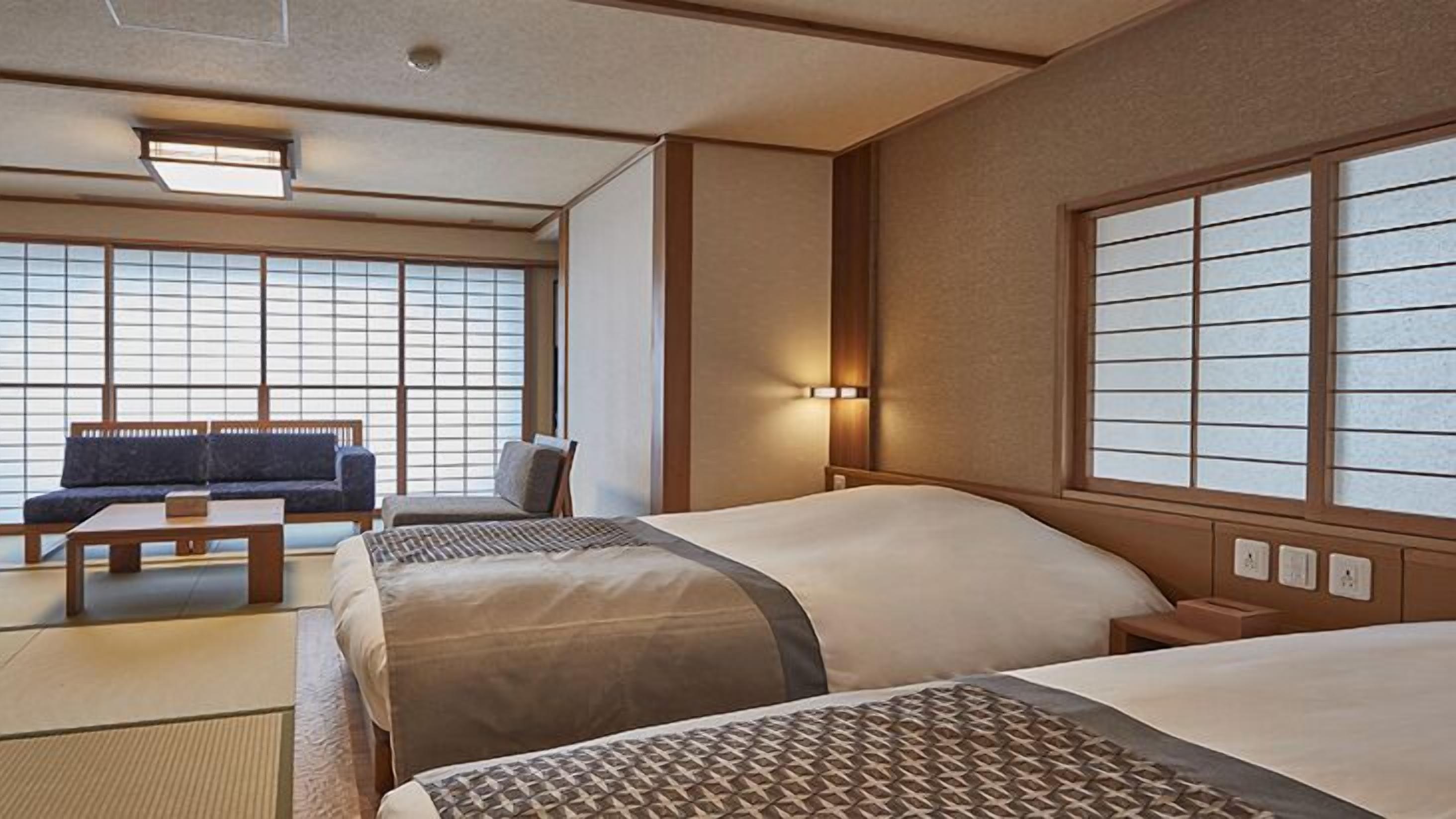 A hotel room with two beds, a sofa, a small table, and large windows with a view.