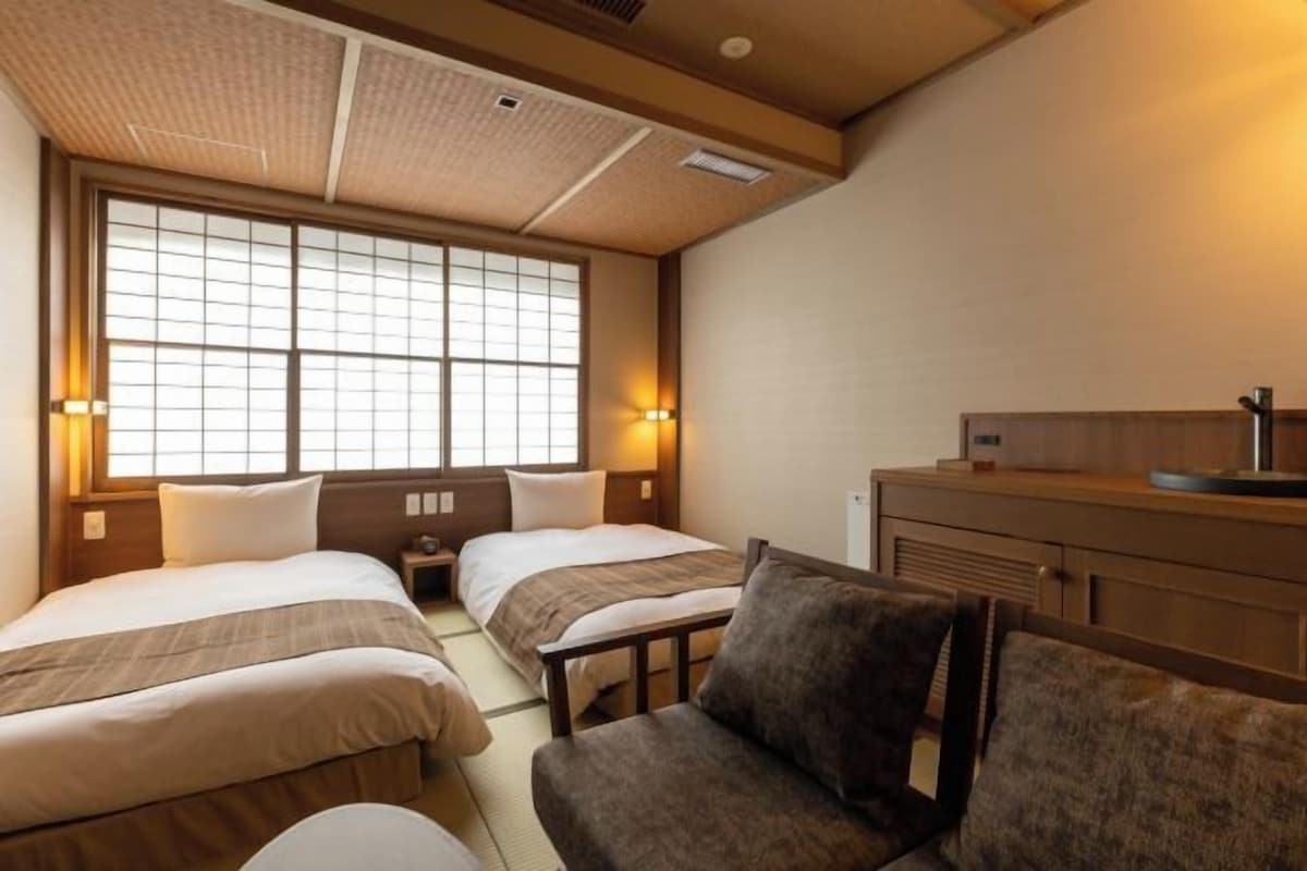 Ein Hotelzimmer mit zwei Betten, einer Couch, einem Schreibtisch und einem großen Fenster mit Jalousien.