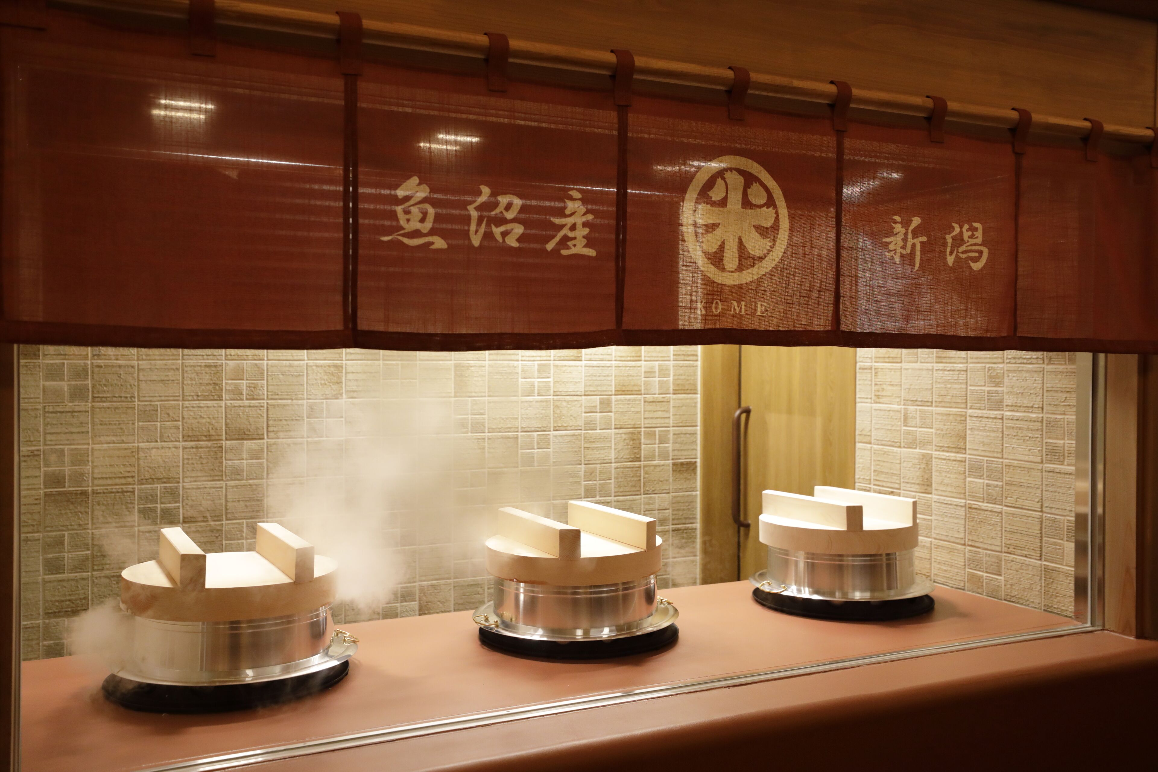 A sushi bar with three wooden blocks on a counter, steam rising from the blocks, and a tiled wall in the background.