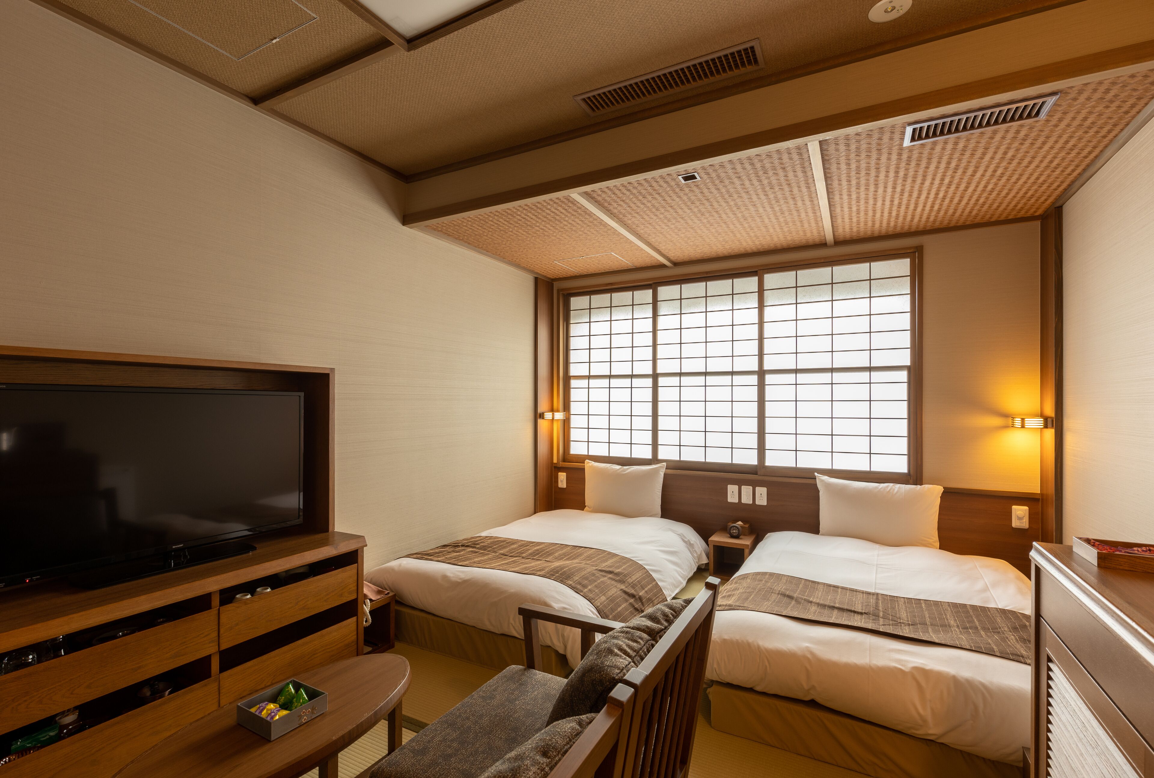 A hotel room with two beds, a TV, wooden furniture, and a large window with blinds.