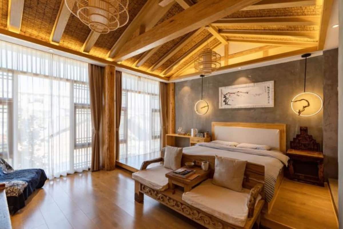 A spacious bedroom with a large bed, a sofa, and a wooden nightstand. There are large windows with curtains, a chandelier, and a wall-mounted artwork.