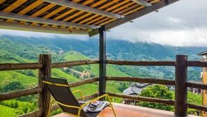 Ein hölzerner Balkon mit einem Stuhl und einer Zeitung, mit Blick auf ein üppiges Tal mit terrassierten Feldern und einem traditionellen Gebäude.