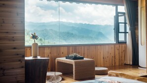 A room with wooden walls, a large window with a mountain view, a small table with a vase and teapot, and two woven poufs. - Floral Hotel Longsheng Yueshangshanshe (Guilin)