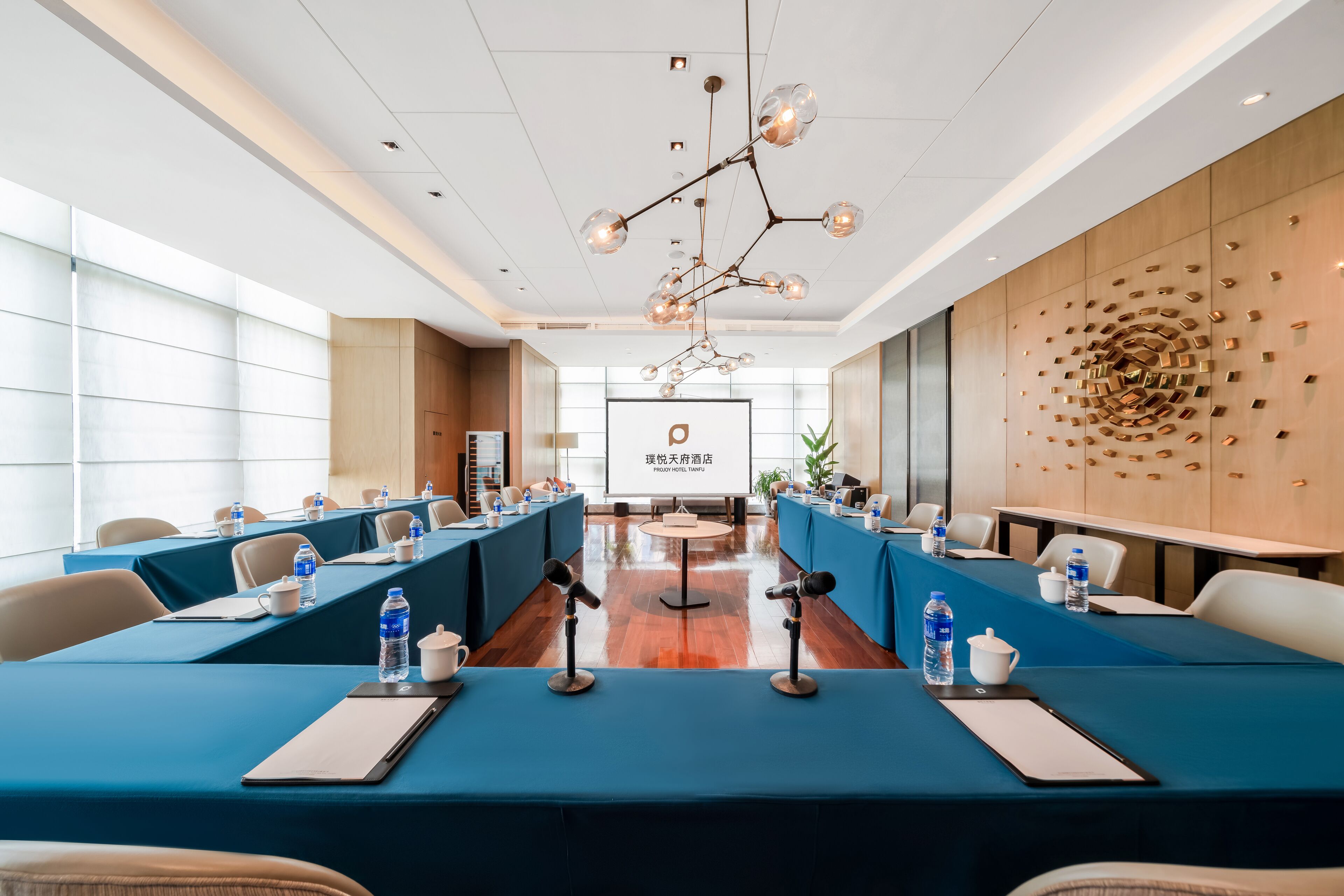 A conference room with a long table, microphones, water bottles, and a projector screen.