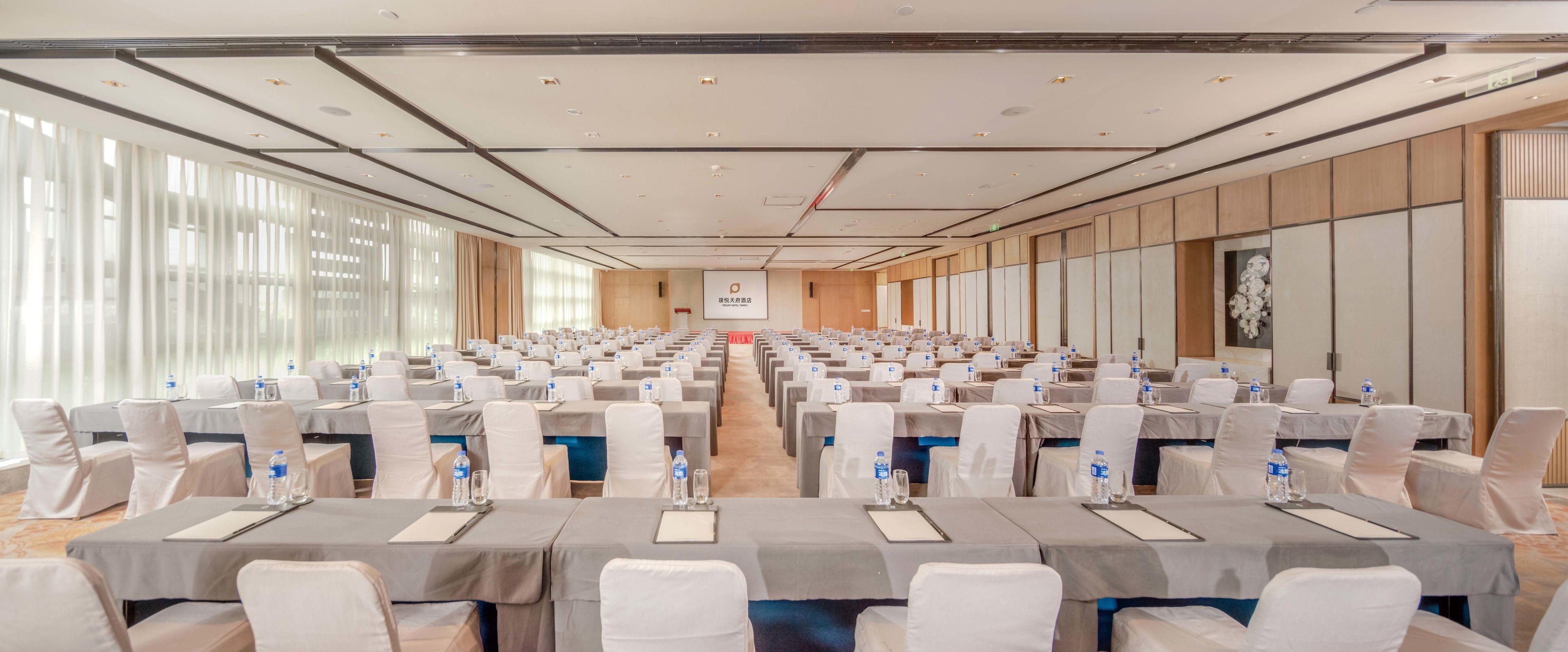A conference room with rows of tables and chairs, large windows, and a wooden panel wall.