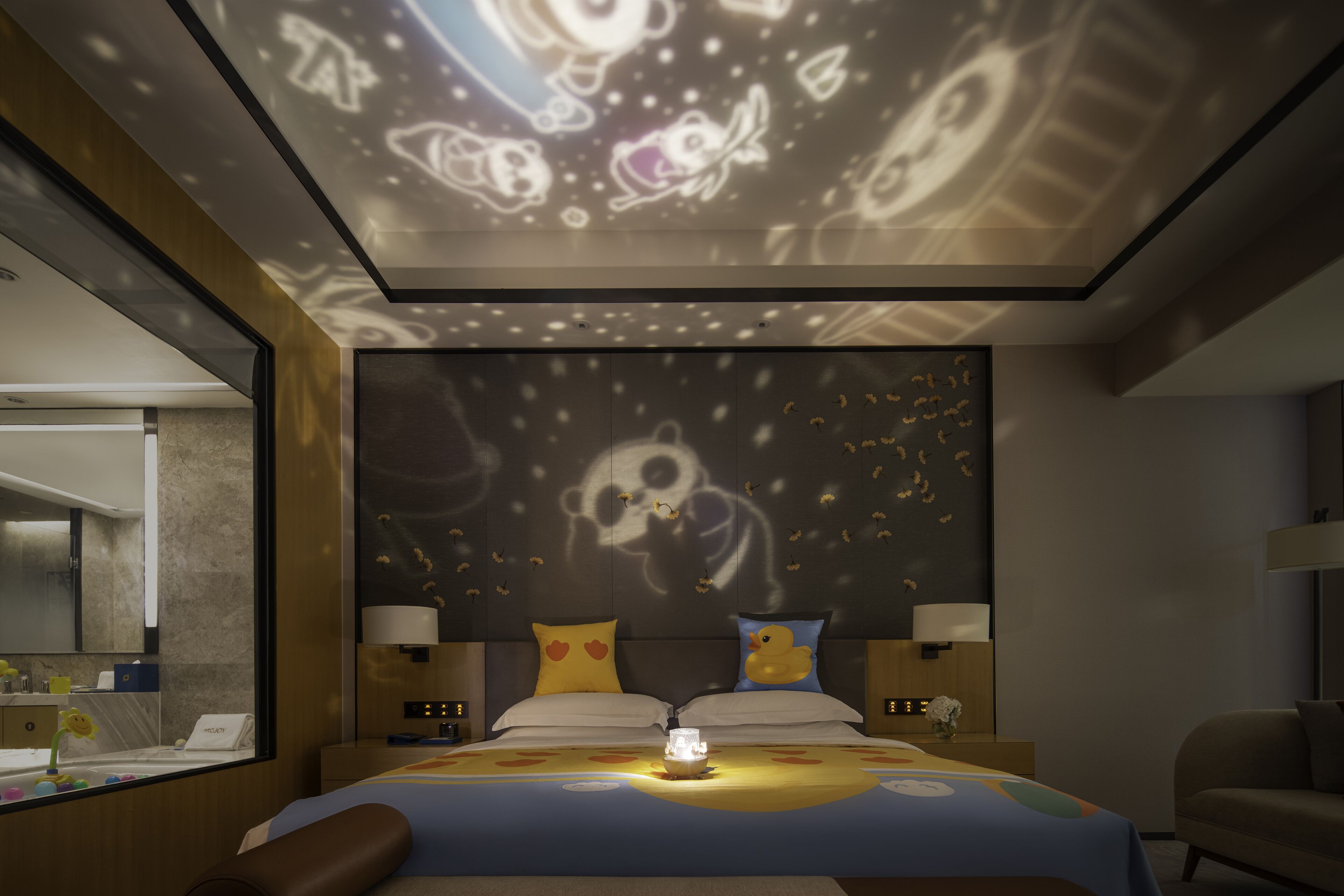 A hotel room with a bed, bedside tables, a nightstand, a lamp, and a ceiling projector displaying animated characters.