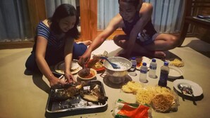 Deux personnes sont assises par terre, préparant un repas à l’aide d’un gril et de différents ustensiles de cuisine.