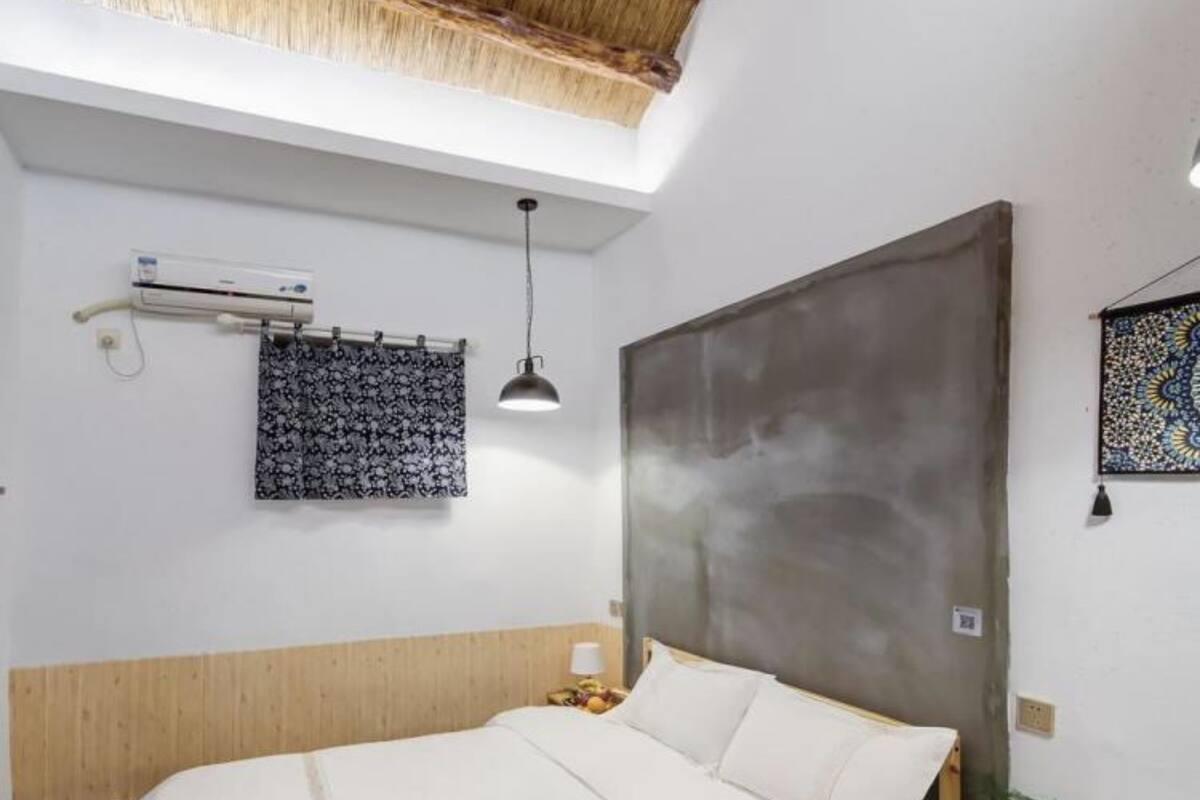 Un dormitorio con una cama grande, cabecera de madera, aire acondicionado instalado en la pared, cortina con estampado, lámpara colgante y una mesita de noche pequeña con una lámpara y una planta.