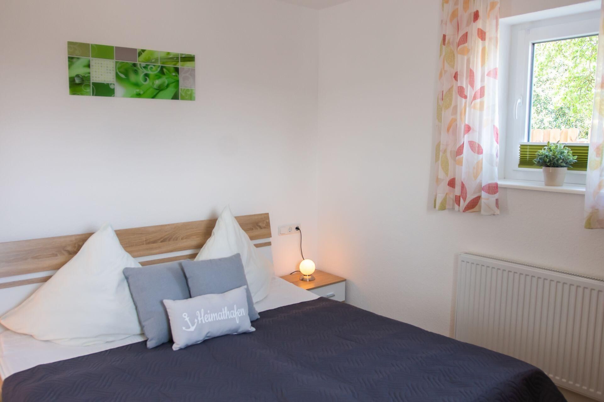A bedroom with a bed, pillows, a bedside table with a lamp, a window with curtains, a radiator, and a wall art piece.