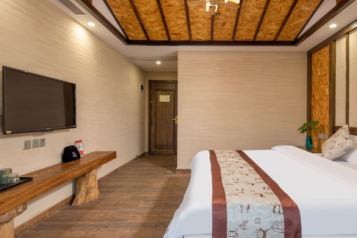 Una habitación de hotel con techo de madera, una cama grande con almohadones estampados, un banco con una botella y un televisor de pantalla plana en la pared.