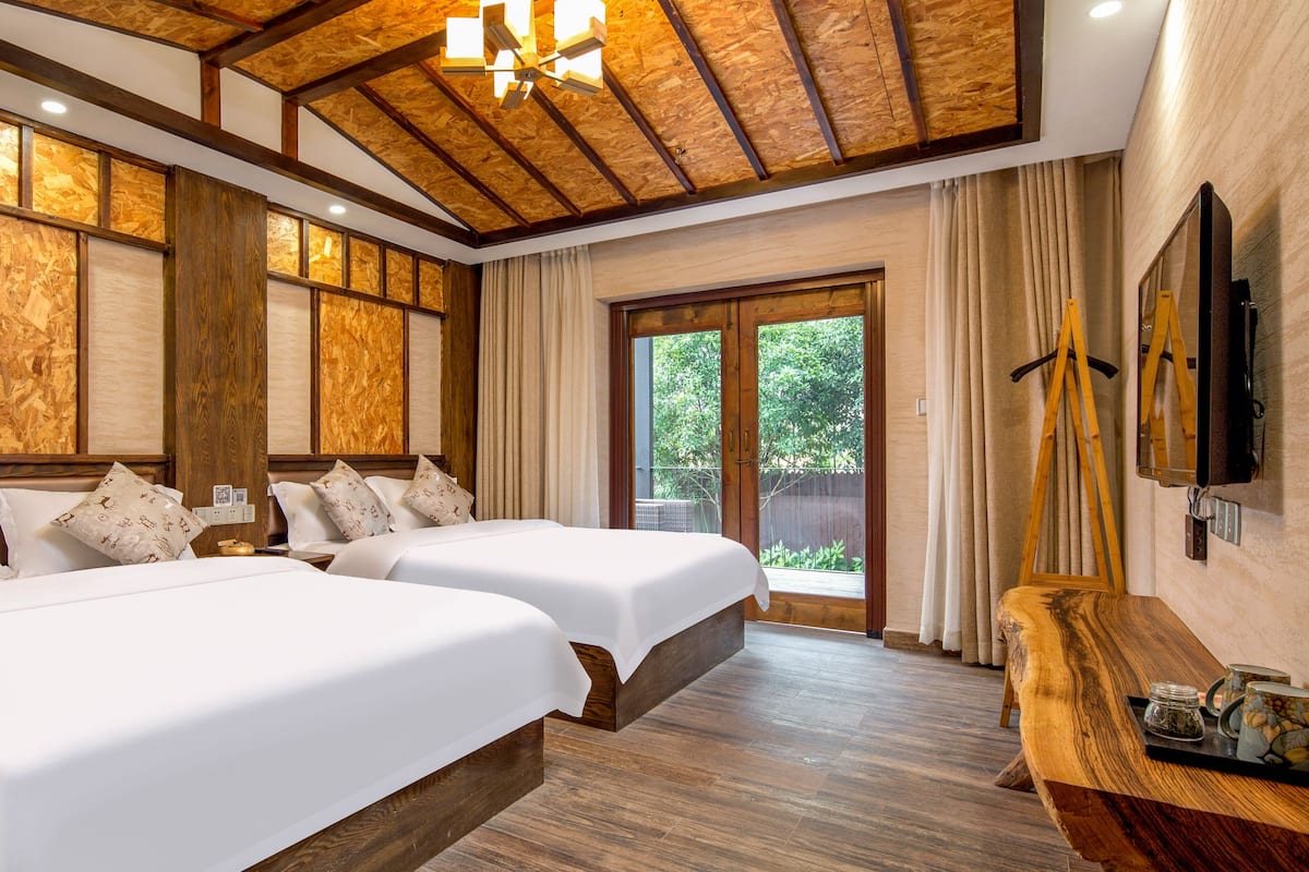 Una habitación de hotel con dos camas, vigas de madera en el techo, un ventanal con cortinas y un aparador de madera con bandeja.