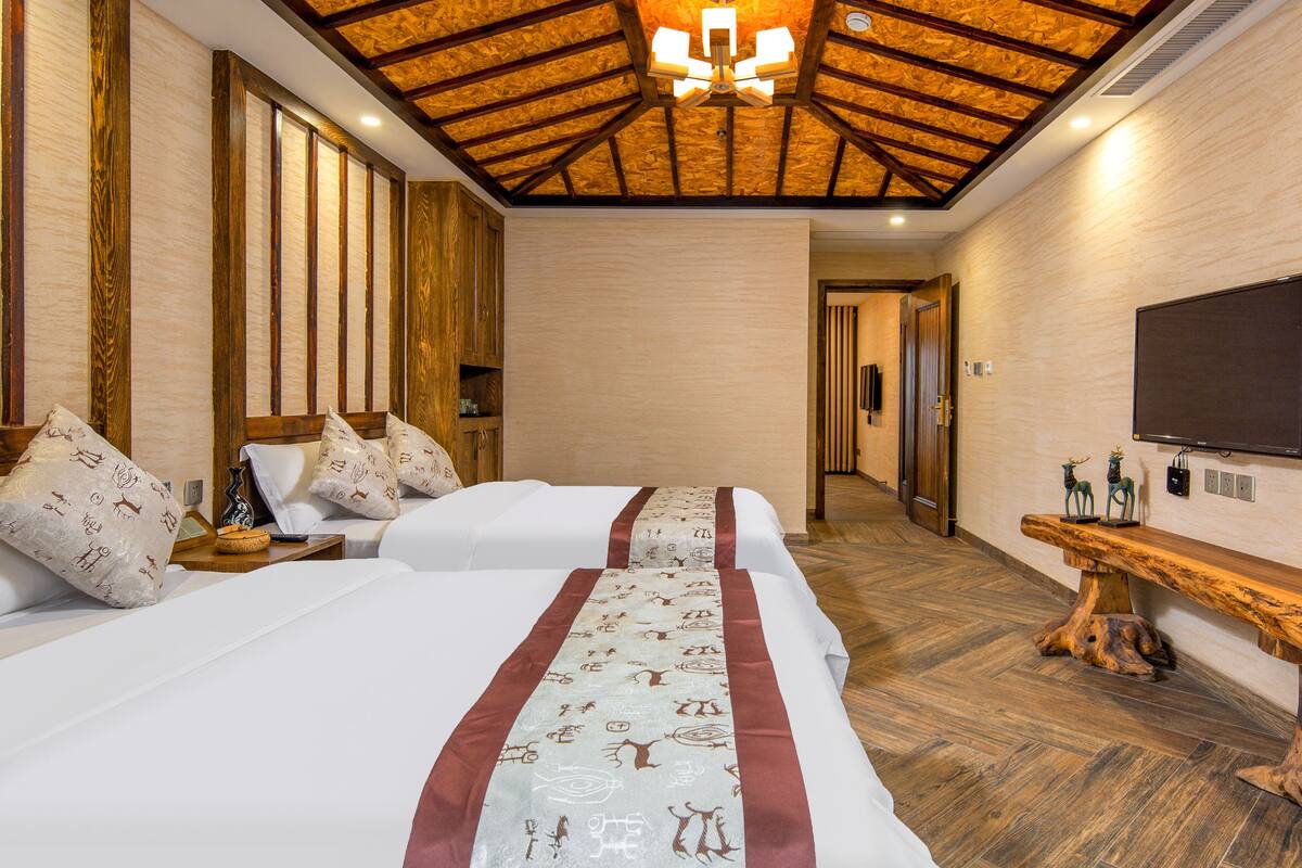 Una habitación de hotel con dos camas, techo de madera, televisión y una banca de madera.