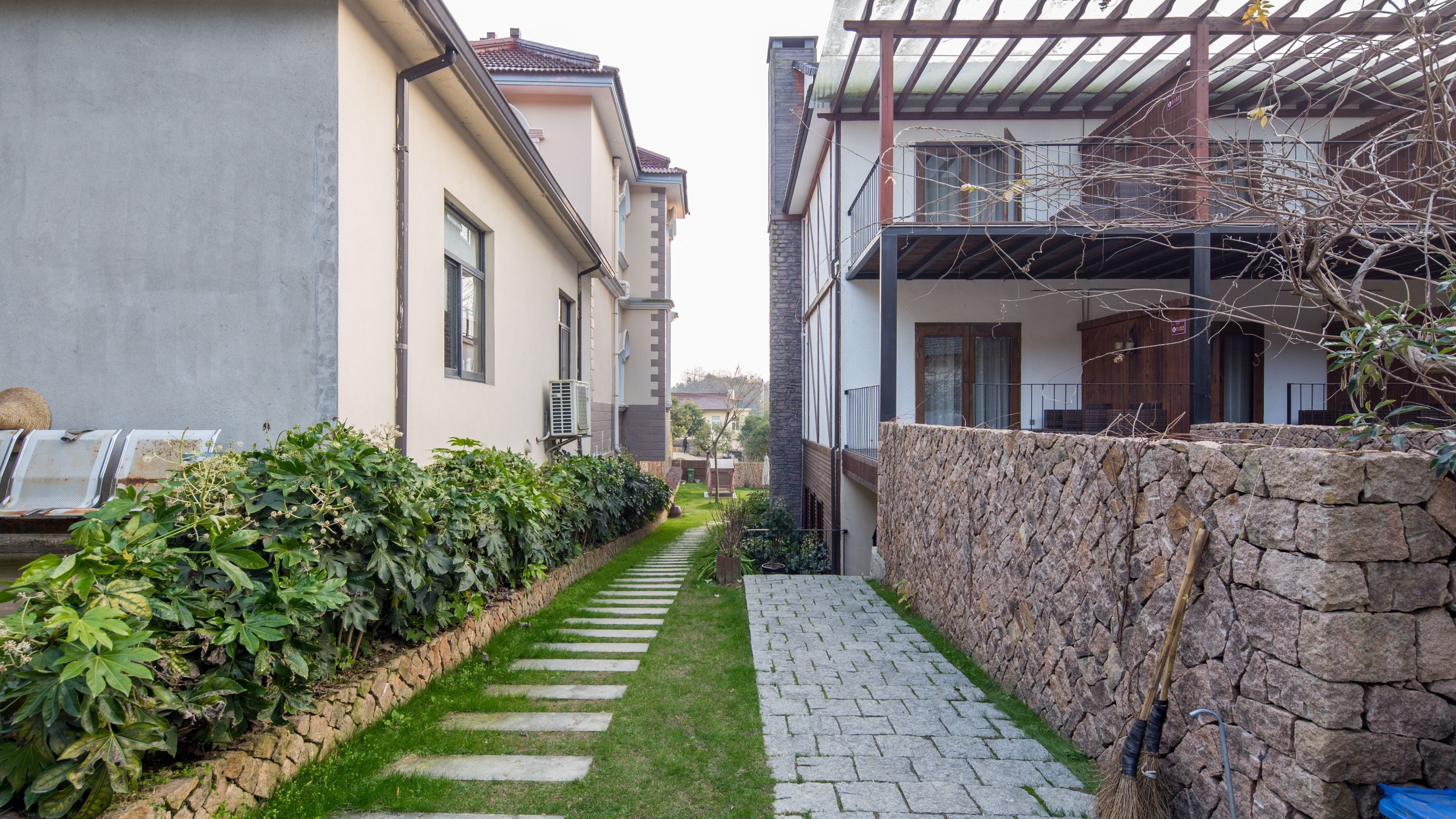 A narrow pathway flanked by stone walls and lush greenery, leading to a building with a stone facade and a covered porch.