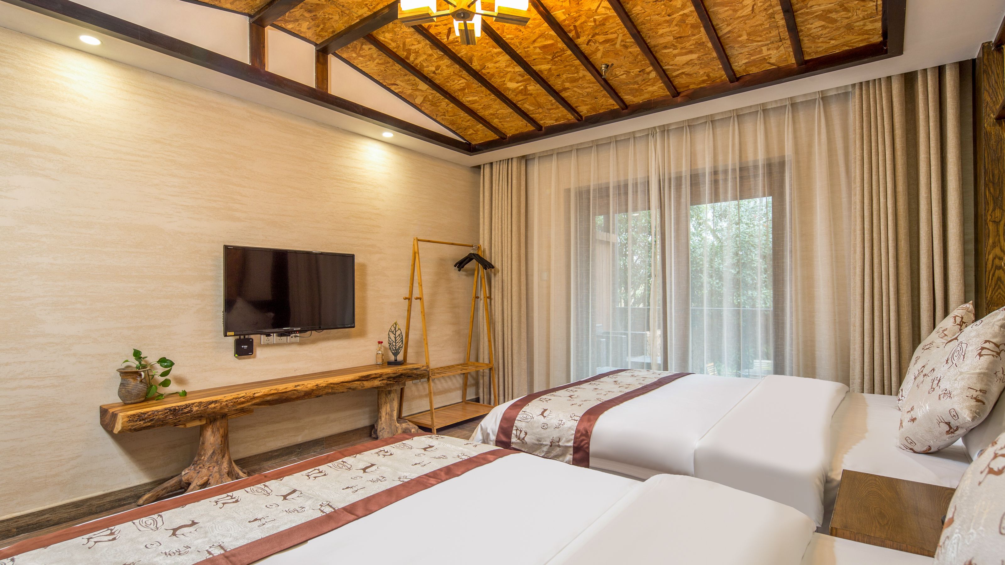 A hotel room with two beds, a TV mounted on the wall, a wooden ceiling, and a large window with curtains.