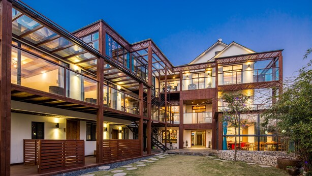 Un bâtiment moderne à plusieurs étages, doté de structures en verre et en bois, d’un espace extérieur aménagé pour s’asseoir et d’un chemin en pierre.