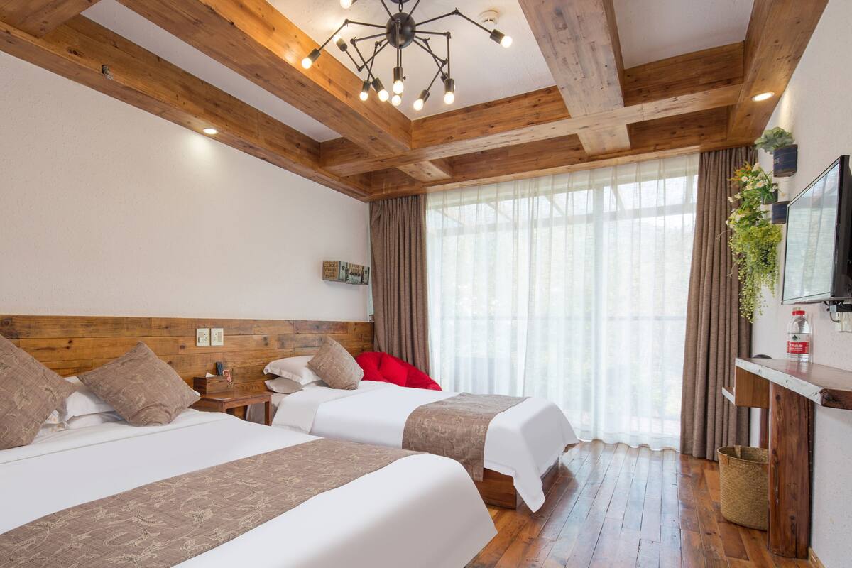 Habitación de hotel con dos camas, vigas de madera, lámpara de techo, ventana con cortinas y un televisor sobre un soporte de madera.