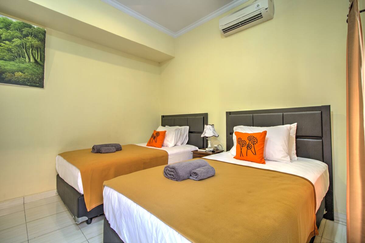 A hotel room with two beds, each with a brown bedspread and white pillows. There is a painting on the wall, a ceiling fan, and an air conditioning unit.