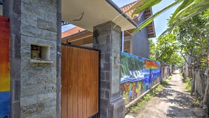 A narrow pathway leading to a house with a colorful mural on the wall, a wooden gate, and a stone pillar. - Nibugi Guest House (Penida Island)