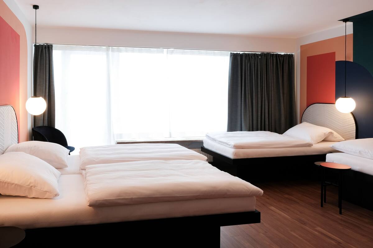 A hotel room with two beds, a small table, and a large window with curtains.
