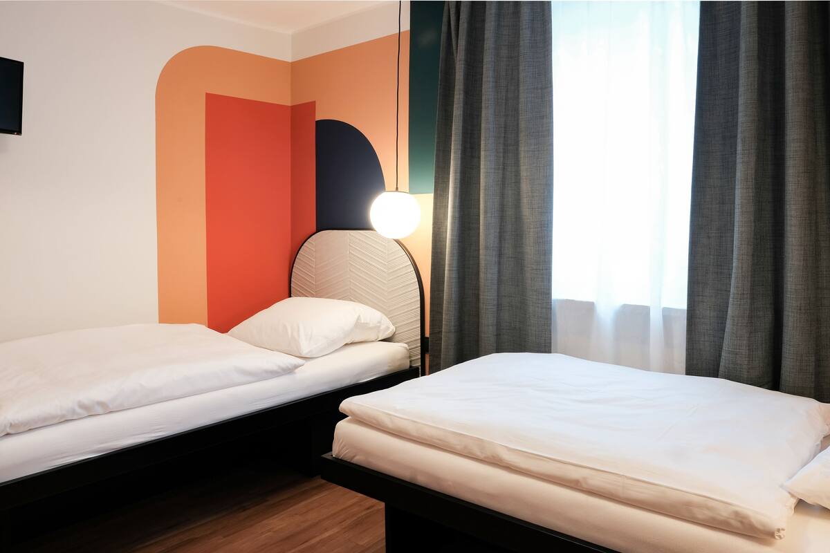 A hotel room with two single beds, a wall-mounted TV, a modern lamp, and a window with curtains.