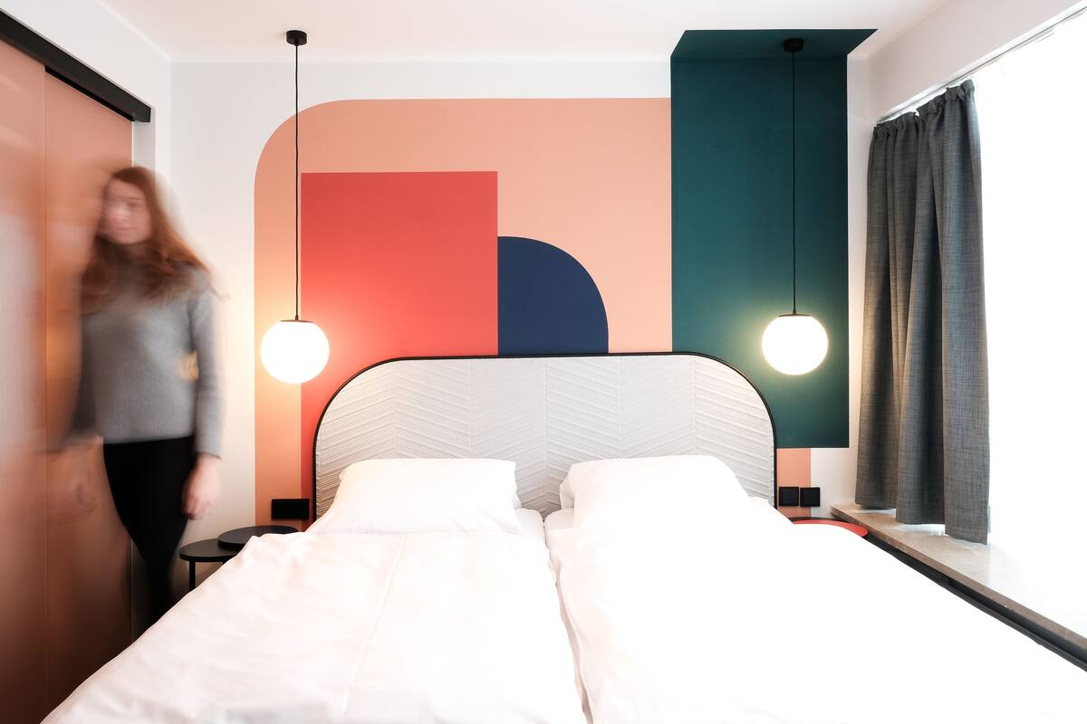 A modern hotel room with a bed, bedside tables, and a large window with curtains.