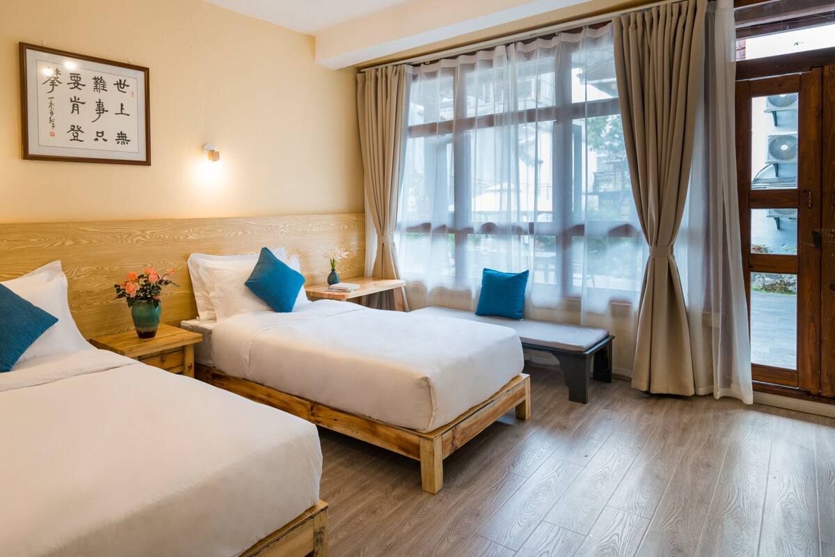 A hotel room with two beds, a wooden headboard, a bedside table with a vase of flowers, a framed calligraphy piece on the wall, and a large window with curtains.